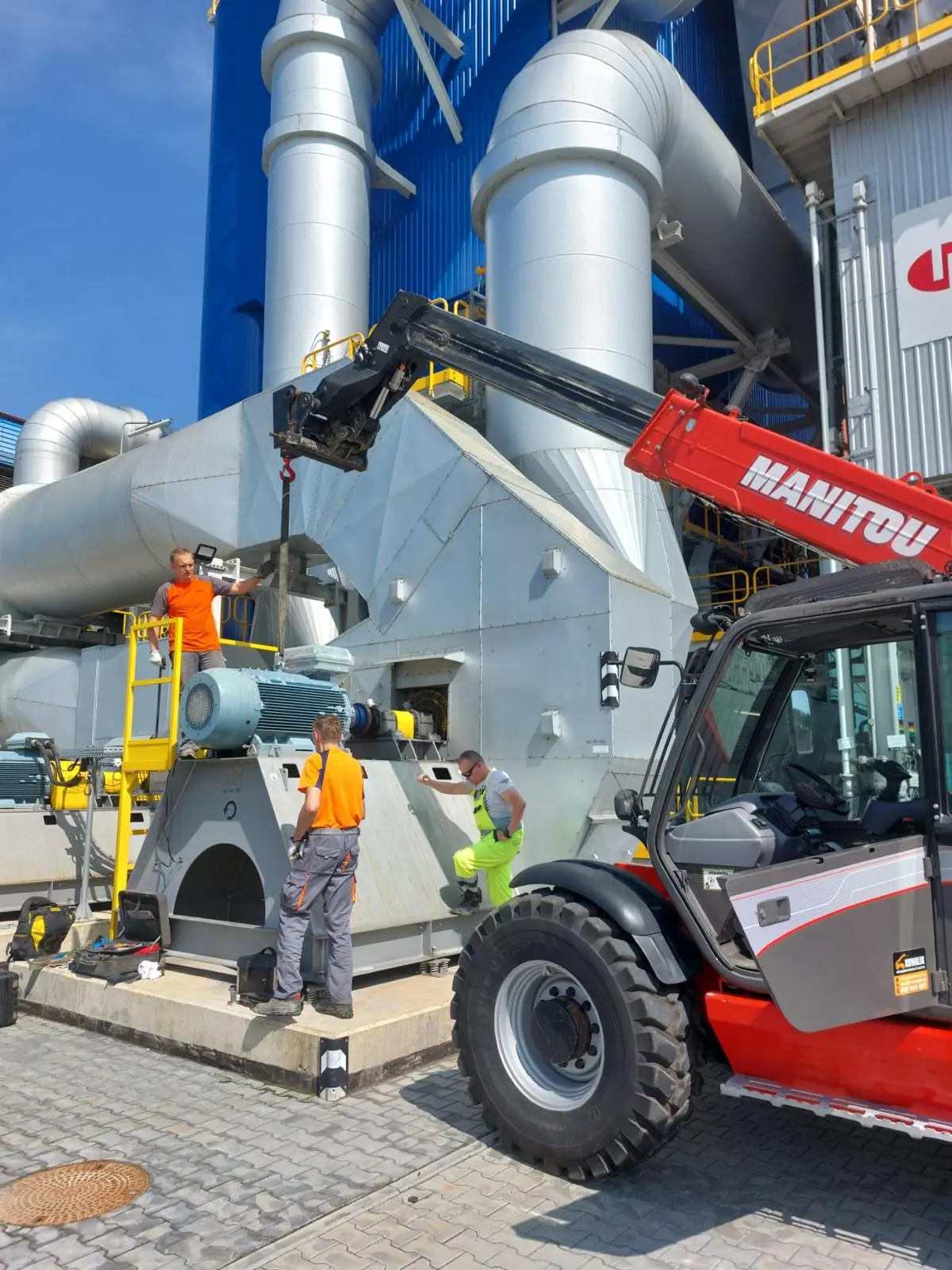 Montaż maszyn przemysłowych – ładowarka teleskopowa Manitou z hakiem podaje element instalacji ekipie montażowej